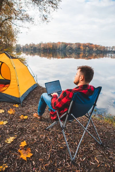 一个人坐在秋湖边的椅子上 研究笔记本电脑自由职业露营的概念 — 图库照片