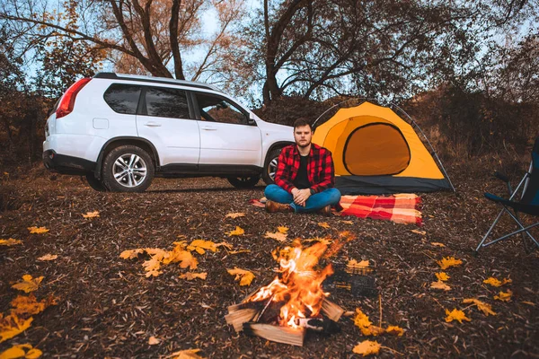 man at camping site sitting near bonfire tent and car on background