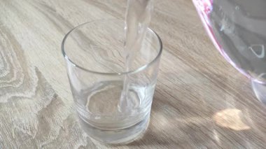 Temiz suyun şeffaf bir sürahide, ahşap bir masada, temiz bir bardağa döküldüğü yakın çekim yatay video. Doğal ışık ve yumuşak sıvı hareketli temiz, ferahlatıcı bir sahne..