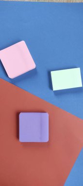 Pembe, yeşil ve mor renkli üç yapışkan nota mavi ve kırmızı bir yüzeye oturur. Kurulum, organize renkler ve malzemelerin yer aldığı bir çalışma alanı sahnesi gösteriyor.