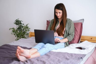 Genç bir kadın dizüstü bilgisayarla yatakta oturuyor. Evde boş zaman.