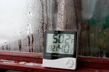 Elektronik saat, takvim, termometre ve higrometre, camdaki yoğunlaşmanın arka planına karşı, yüksek nem oranı. Laboratuvardaki dijital higrometre farenjitin sıcaklığını gösteriyor.
