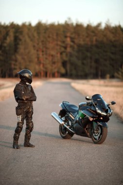 Deri kıyafetli motosikletçi ve kask yolda durur ve bir spor motosikletine bakar.
