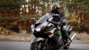 Yakışıklı motosikletçi deri ceket giyiyor ve yolda kask tutuyor.