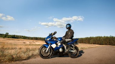 Deri ceketli, kasklı motosikletli bir adam yola bakıyor.