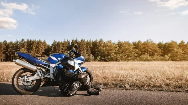 Deri kıyafetli ve siyah kasklı bir bisikletçi asfalt yolda bir spor motosikletinin yanında oturur ve fotokopi çekerek uzaklığa bakar.