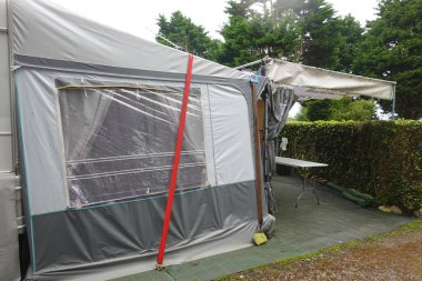 Gri karavan tentesi, çitlerle ve yeşil çimlerle çevrili bir masanın bulunduğu bir kamp alanında kırmızı kayışla korunuyor.