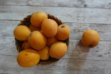 Hasır bir sepetten taşan olgun lokuatlar, rustik ahşap bir masanın üstünde canlı bir ekran oluşturuyor.