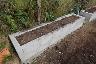 Beton blok yapısı, yeni sebze ve bitkiler ekmeye hazır, zengin toprakla dolu, yükseltilmiş bir bahçe yatağı oluşturuyor.