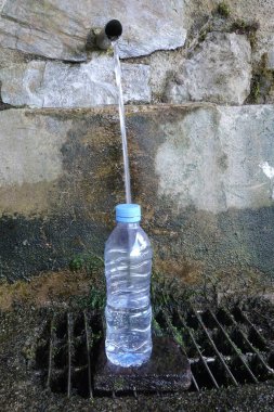 Metal bir borudan şeffaf bir plastik şişeyi dolduran temiz doğal su akıntısı yosunlu bir drenaja doğru akıyor.