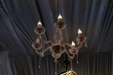 Parlak mum benzeri ampuller içeren güzel ve süslü bir kristal avize yumuşak örtülü koyu kumaş tavandan zarif bir şekilde sarkıyor, lüks ve atmosferik iç tasarım elementi yaratıyor..