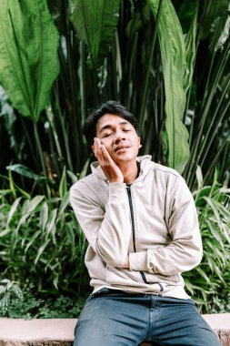 Gözleri kapalı sakin bir genç adam elini yüzüne koyar, yemyeşil tropikal yaprakların arasında rahat bir şekilde oturur. Doğadaki sakinlik, rahatlama ve farkındalığı somutlaştırır..