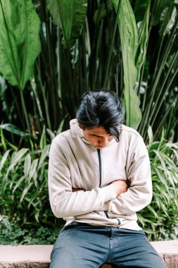 Genç bir adam dışarıda oturuyor, kollarını karnına kavuşturmuş, karın rahatsızlığı ya da derin düşünceler yaşıyor gibi görünüyor. Etrafı canlı yeşil tropikal yapraklarla çevrili..