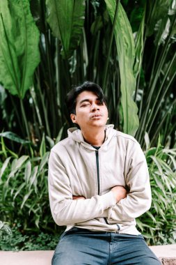 Genç Asyalı bir adam gözleri kapalı ve kolları çapraz bir şekilde canlı yeşil bir bahçede sakin bir anın tadını çıkararak huzur içinde oturur. Rahat bir açık hava yaşam tarzını yansıtan sıradan bir kapüşonlu giyiyor..