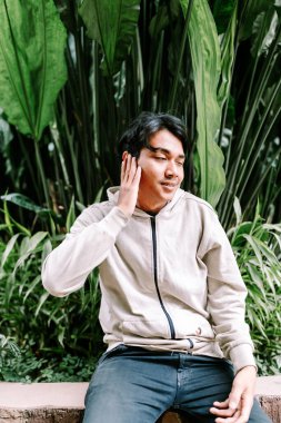 Bej kapüşonlu neşeli genç bir adam kulağına dokunuyor, açık havada yemyeşil tropikal yaprakların arasında nazikçe gülümsüyor. Doğada rahat ve hoşnut görünüyor..