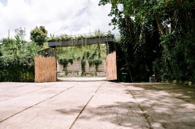Açık bir bambu kapısının geniş açılı görüntüsü ziyaretçileri canlı, fazla gelişmiş bir tropikal bahçeye davet ediyor. Bulutlu bir gökyüzünün altındaki yeşilliğe doğru uzanan beton bir yol var..