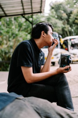 Düşünceli, genç Asyalı bir adam sıradan bir ortamda oturuyor, elinde bir kamerayla, dalgın dalgın bakıyor..