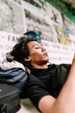 Genç bir adam umumi bir alanda mavi bir sırt çantasının üzerinde dinleniyor, dinleniyor ya da uyuyor. Bu yolculuk sırasında yorgunluğu ya da dinlenmeyi gösterir..