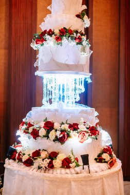 Göz kamaştırıcı çok katlı bir düğün pastası güzel kırmızı, beyaz ve pembe güllerle süslenmiş, narin fırfırlar ve parlak bir kristal avize aydınlatma armatürü. Zarif bir kutlama için mükemmel.