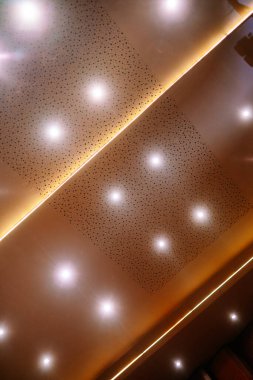 Parlak spot ışıkları ve çarpıcı çapraz LED ışık şeridine sahip çağdaş bir tavanın soyut görüntüsü. Delinmiş akustik paneller bu mimariye modern bir doku ekler.