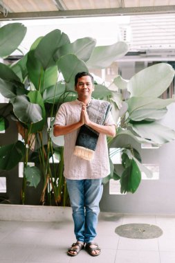 Bir adam, elleri geleneksel bir saygı göstergesi ya da selamlama jesti ile, yemyeşil tropikal bitkilerin arka planına karşı katlanmış bir dua paspasını tutuyor..