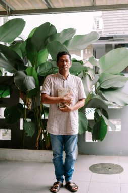 Gözleri kapalı bir adam, dindar bir kitabı göğsüne dayayarak bir anlık bağlılık içinde ayakta duruyor. Sıradan giyinmiş, büyük tropikal bitkiler ve dingin bir duvar örüyor.