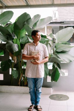 Yetişkin bir adam açık havada durup göğsüne süslü bir kitap tutuyor. Sıradan giyinmiş ve yemyeşil tropikal bitkilerin önüne yerleştirilmiş.