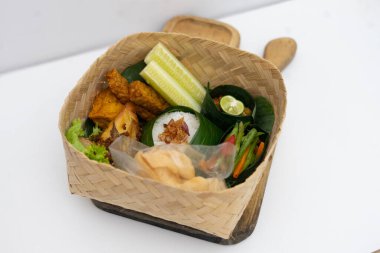 Muz yaprağına sarılı pirinç, kızarmış tofu, sebze ve sambal içeren otantik bir Güneydoğu Asya yemeği, temiz beyaz arka planda geleneksel bir bambu sepetinde servis ediliyor..