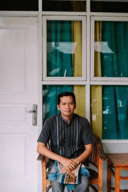 Sakin bir ifadeye sahip yetişkin bir adam ahşap bir sandalyeye oturmuş, geleneksel desenli bir gömlek giyiyor ve elinde dekoratif bir bez tutuyor. Arka planda penceresi olan bir evde oturuyor..