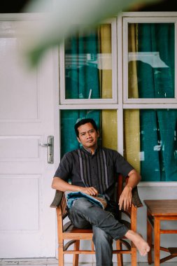 Huzurlu bir Asyalı adam geleneksel ahşap bir sandalyede, desenli bir gömlek ve kot pantolon giyer. Evde rahat görünüyor, doğrudan izleyiciye bakıyor..