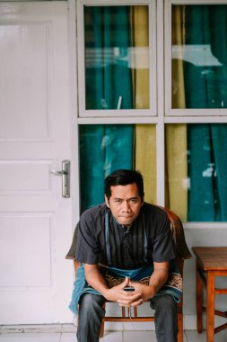 Dalgın bir Asyalı adam sakince kapalı bir yerdeki tahta sandalyeye oturur ve doğrudan kameraya bakar. Geleneksel desenli gömleği ve sarong benzeri kumaşı kültürel kıyafetleri yansıtıyor..