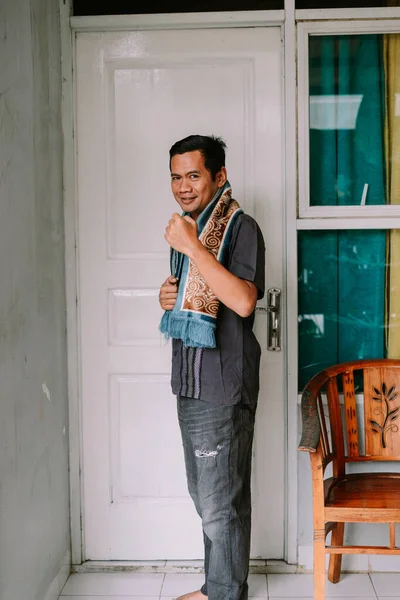 Dost canlısı Asyalı bir adam kameraya gülümsüyor, canlı desenli bir eşarp ve günlük giysiler giyiyor, beyaz bir kapının yanında duruyor..