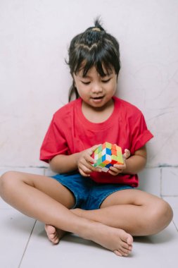 Genç bir çocuk, rengarenk bir bulmaca küpünü manipüle etmeye odaklanmış bir şekilde yerde oturuyor. Bu resim erken çocukluk gelişimini, bilişsel becerileri ve oyun yoluyla problem çözmeyi temsil eder..