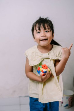 Neşeli bir Asyalı genç kız renkli bir bulmaca küpü tutuyor ve yukarı doğru işaret ediyor, coşku ve parlak bir fikir. Bu an çocukluk eğitimini ve eğlenceli keşfi yakalar..