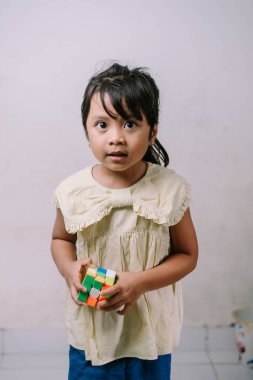 Meraklı küçük bir kız canlı, renkli bir bulmaca küpünü elinde tutuyor ve doğrudan kameraya bakıyor. Bu resim çocukluk masumiyetini, öğrenmeyi ve eğlenceli problem çözmeyi yakalar..