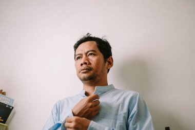 Açık mavi yakalı, yetişkin bir adam kollarını düzeltir. Bakışları düşünceli ve yukarı yönlüdür. Basit bir arkaplan onun odaklanmış ifadesini vurgular, hazırlığı veya