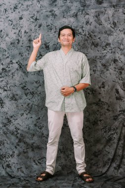 Neşeli Asyalı bir adam günlük giysiler içinde gri bir zemine karşı duruyor, kameraya bakarken mutlu bir gülümsemeyle parmağını yukarı kaldırıyor..