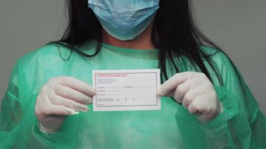 Koruyucu yeşil takım elbiseli kimyagerin elinde covid-19 aşı kartı var. Yüz maskesi ve beyaz eldivenli bir kadının Coronavirus koruma aşısı için belge göstermesi..