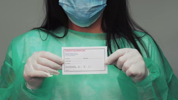 緑のスーツを着た化学者は Covid 19予防接種カードを保持しています 顔マスクと白い手袋を持つ女性の医療概念は コロナウイルス保護予防接種ショットのための文書を表示します — ストック動画