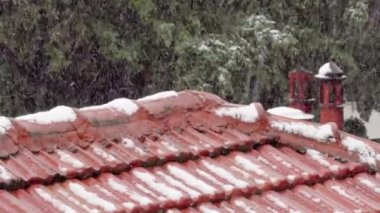 Etrafı ağaçlarla çevrili seramik kil çatıya kar yağıyor. Şiddetli kar yağışlı kış manzarası, üst üste binen kırmızı kiremitler üzerine beyaz pullar düşüyor..