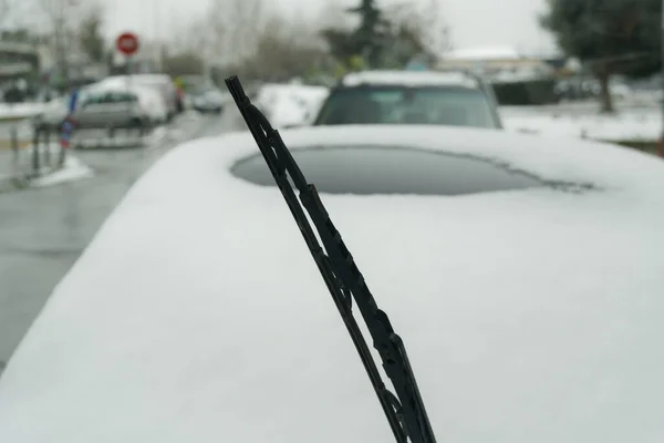 Lifted screen wiper before a car covered with snow. Day view of frozen windshield blade raised up against blurred background of a parked vehicle on a cold winter day.