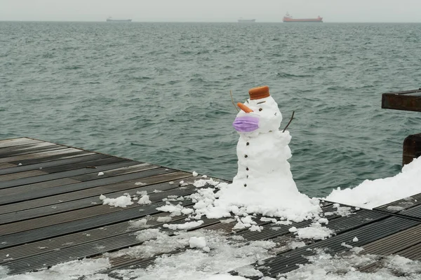 Funny snowman with face protection mask by the waterfront during the coronavirus pandemic. Day view of cute white frozen construction with covid-19 mask and carrot nose against sea background.