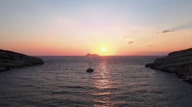 Ufukta ada görünmeden önce küçük bir yolcu gemisi insansız hava aracı ile turuncu gün batımı. Girit Adası 'ndaki Matala kireçtaşı mağaralarının sakin sulardaki tekneleriyle kıyı üzerinde dönen hava akşam manzarası..