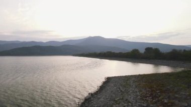 Çakıl taşlı rıhtıma uzanan yeşil ağaçlı bir gölün havası. Kuzey Yunanistan 'daki Kerkini su sahasının arka plan dağ manzaralı insansız hava aracı görüntüsü. 