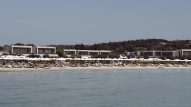 Chalkidiki, Yunanistan - 26 Haziran 2021: Şemsiyeli deniz yatakları üzerinde plaj banyosu yapan modern deniz manzarası. Kassandra Chalkidiki yarımadasında yeşil bitki örtüsüyle çevrili Sani lüks tatil yeri..