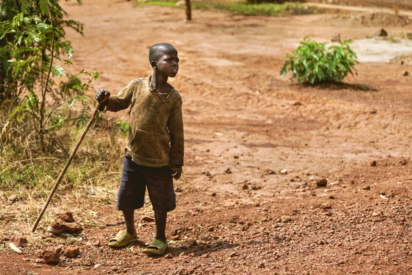 O menino fica na estrada da terra com suas roupas velhas e rasgadas . — Fotografia de Stock