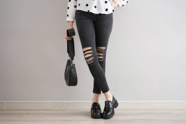 Gri arka planda çantası ve ayakkabısı olan kot pantolonlu genç bir kızın bacakları..