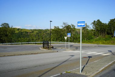 Norveç 'te sakin bir banliyö yolunda yaya geçidi ve otobüs durağı işaretleri. Yol işaretleri, trafik işaretleri ve açık mavi gökyüzünün altında yeşil doğal manzarası olan boş bir cadde. Yol güvenliği kavramı, toplu taşıma, altyapı ve susta