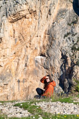 Turuncu ceketli genç bir kadın yüzünü kameraya çevirir ve kendini El Chorro, Ardales, İspanya 'da güneşten korumak için eliyle yüzünü kapatır. Bulanık kayalık duvar arkaplanı, macera ve doğa tarzıyla bulanık yakın plan.