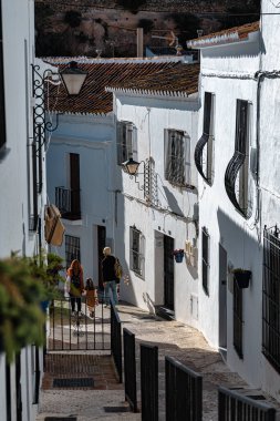 Tipik Endülüs kıvrımlı ara sokakları, 21 Şubat 2026 'da Mijas, Endülüs' te beyaz cepheler, siyah korkuluklar ve demir korkuluklar. Bir çift gerçek bir seyahat sahnesinde küçük bir kızla kameraya doğru yürür..